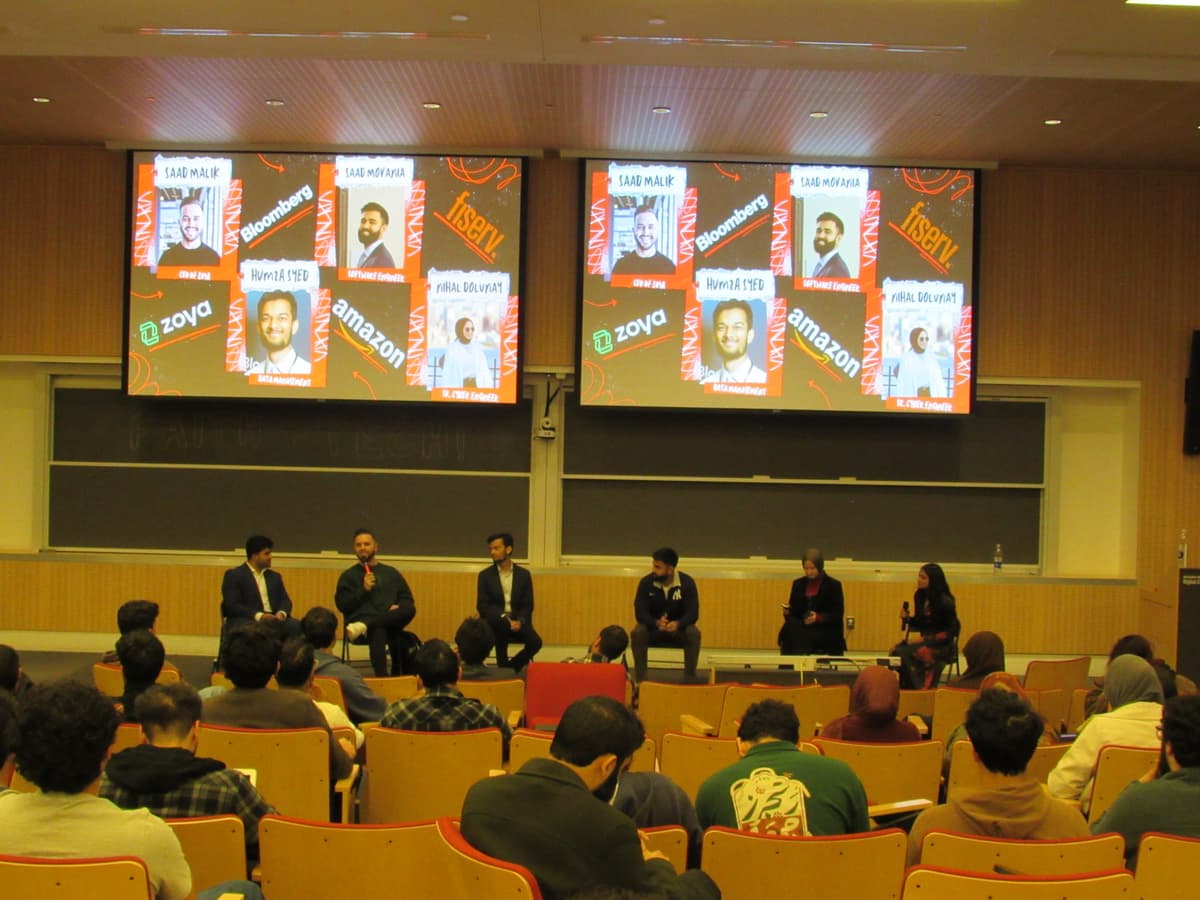 Wide view of the full panel, five speakers on stage, packed audience, dual screens